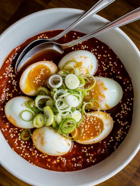 Overhead view of six soft boiled eggs sitting in a deep red spicy gochujang sauce in a white ceramic bowl, eggs lightly stained from the sauce, topped generously with sliced green onions and sesame seeds with two silver spoons resting in the bowl