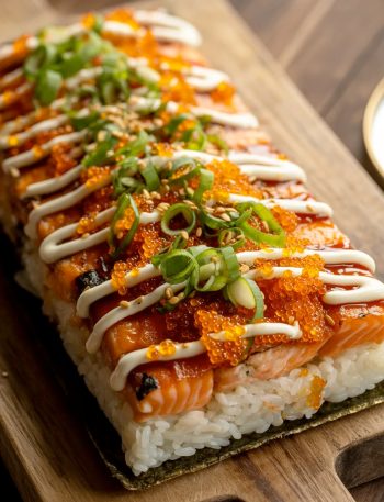 A rectangular spicy salmon sushi bake on a wooden serving board showing a caramelized golden salmon topping covered in bright orange tobiko fresh green onions sesame seeds and herbs sitting on a compact white sushi rice base