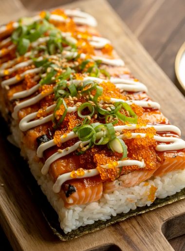 A rectangular spicy salmon sushi bake on a wooden serving board showing a caramelized golden salmon topping covered in bright orange tobiko fresh green onions sesame seeds and herbs sitting on a compact white sushi rice base