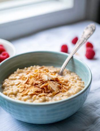 A light blue bowl of creamy coconut oats topped with golden toasted coconut flakes and a silver spoon on a light grey surface