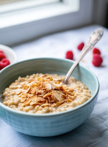 A light blue bowl of creamy coconut oats topped with golden toasted coconut flakes and a silver spoon on a light grey surface