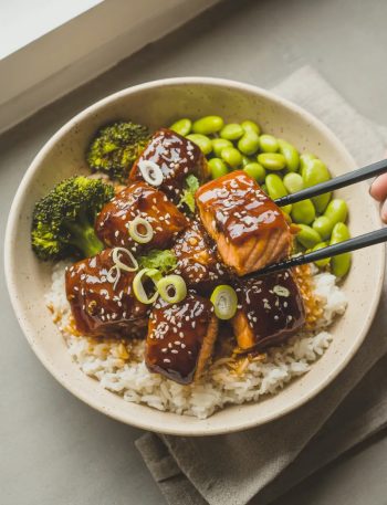 Teriyaki glazed salmon bites over white rice in a wide ceramic bowl with steamed broccoli edamame sliced green onions and sesame seeds with black chopsticks lifting one piece