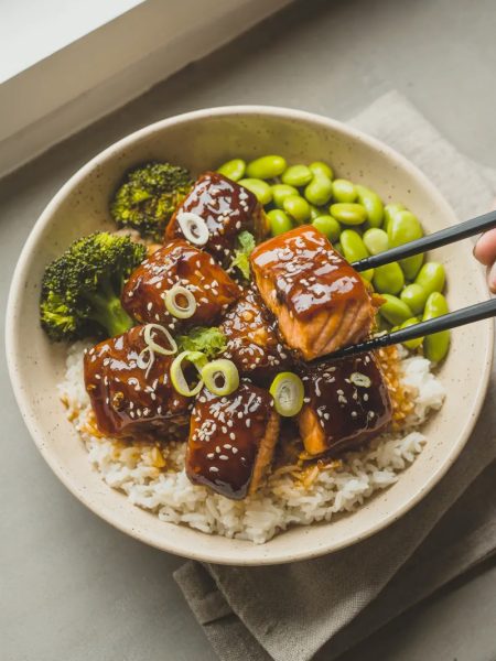 Teriyaki glazed salmon bites over white rice in a wide ceramic bowl with steamed broccoli edamame sliced green onions and sesame seeds with black chopsticks lifting one piece