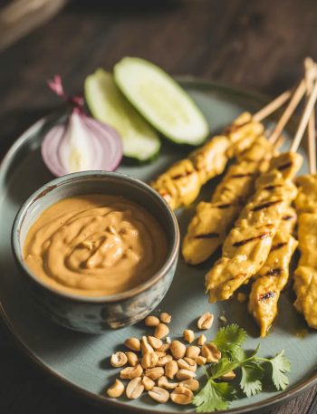 Thai chicken satay skewers served on a black plate with a bowl of homemade peanut dipping sauce garnished with crushed peanuts and fresh cilantro