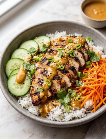 A stone bowl filled with fluffy white rice topped with sliced grilled chicken cucumber rounds shredded carrots and a generous drizzle of creamy Thai peanut sauce garnished with green onions and sesame seeds