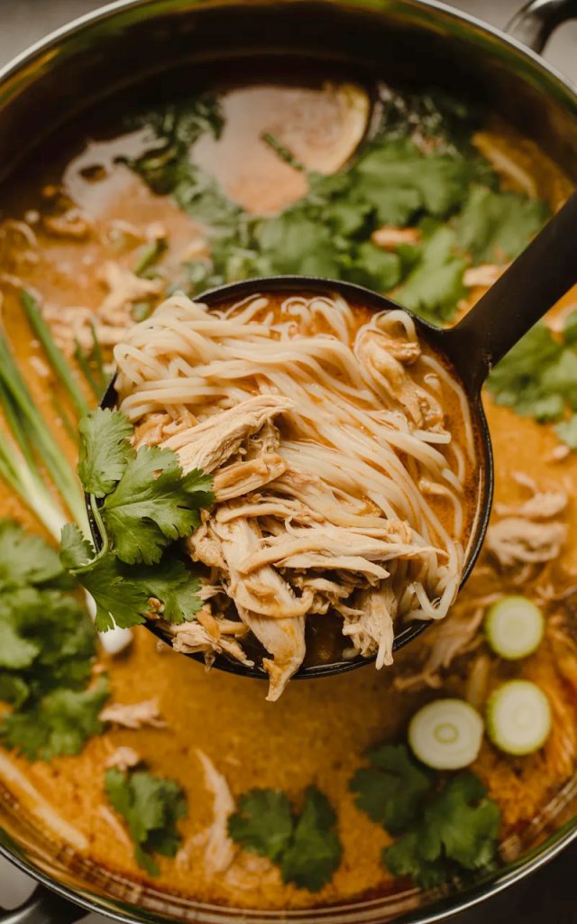 A black ladle lifting silky rice noodles and tender shredded chicken from a rich golden coconut curry broth in a large pot garnished with fresh cilantro