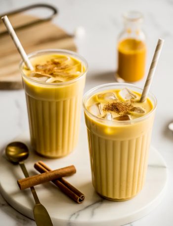 Two clear plastic cups filled with creamy iced golden milk turmeric lattes with straws and a light cinnamon dusting on top sitting on a white marble surface with cinnamon sticks and a gold spoon