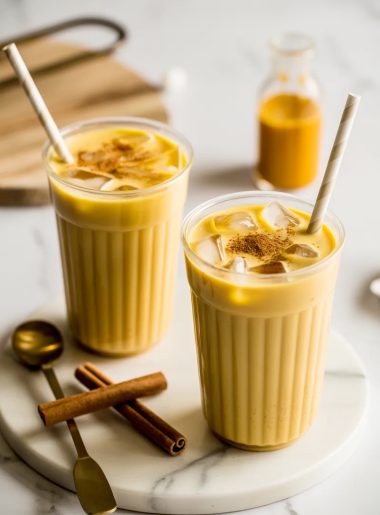 Two clear plastic cups filled with creamy iced golden milk turmeric lattes with straws and a light cinnamon dusting on top sitting on a white marble surface with cinnamon sticks and a gold spoon