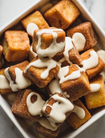 Overhead close-up of a white square bowl filled with golden cinnamon sugar angel cake churro bites drizzled with white vanilla glaze showing their crispy caramelized exterior
