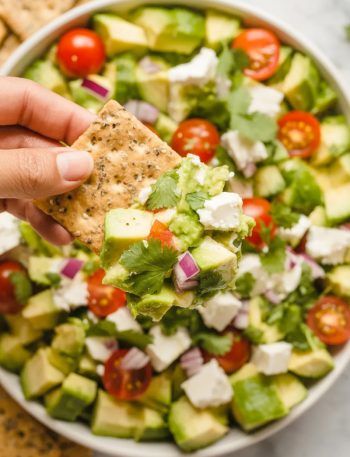 Overhead close-up of a white bowl filled with vibrant avocado feta dip showing diced avocado crumbled feta quartered cherry tomatoes diced cucumber red onion and fresh herbs with a hand holding a cracker loaded with dip above the bowl