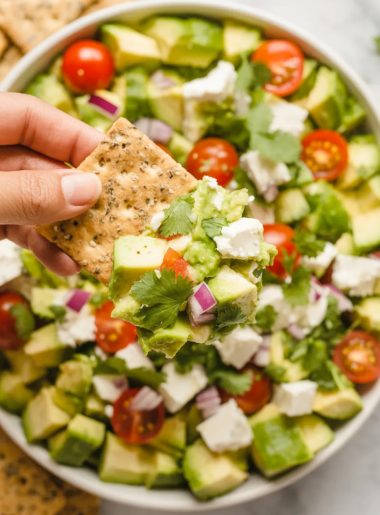 Overhead close-up of a white bowl filled with vibrant avocado feta dip showing diced avocado crumbled feta quartered cherry tomatoes diced cucumber red onion and fresh herbs with a hand holding a cracker loaded with dip above the bowl