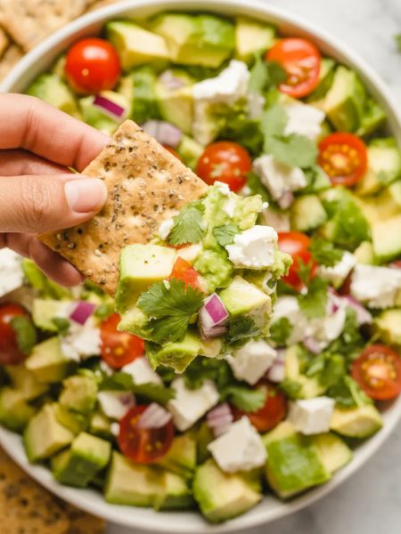 Overhead close-up of a white bowl filled with vibrant avocado feta dip showing diced avocado crumbled feta quartered cherry tomatoes diced cucumber red onion and fresh herbs with a hand holding a cracker loaded with dip above the bowl