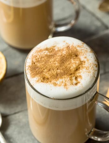 Close-up overhead shot of a tall clear glass mug filled with a creamy chai tea latte showing a thick layer of white steamed milk foam on top dusted with ground cinnamon on a gray stone surface