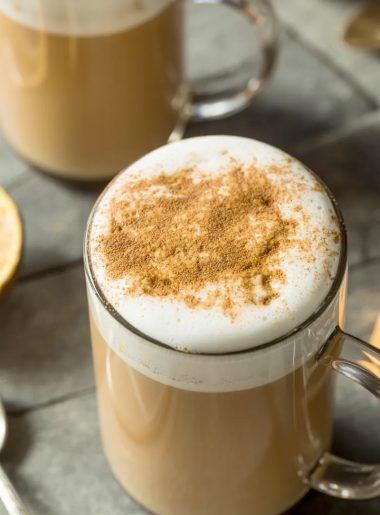 Close-up overhead shot of a tall clear glass mug filled with a creamy chai tea latte showing a thick layer of white steamed milk foam on top dusted with ground cinnamon on a gray stone surface