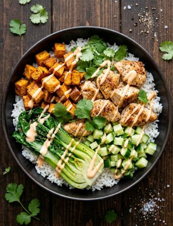 Chicken and sweet potato bowls with roasted sweet potatoes, seared chicken, fresh cucumber, wilted greens, white rice and creamy sriracha sauce in a ceramic bowl