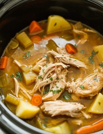 Overhead view of a crockpot filled with tender shredded chicken thigh pot roast with carrots potatoes celery and rich savory broth garnished with fresh parsley