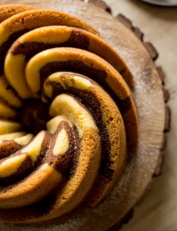 Overhead close-up of a fully baked golden cinnamon swirl bundt cake on a wooden cake stand showing the dramatic dark cinnamon sugar swirl pattern spiraling through the ridged golden cake
