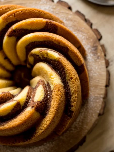 Overhead close-up of a fully baked golden cinnamon swirl bundt cake on a wooden cake stand showing the dramatic dark cinnamon sugar swirl pattern spiraling through the ridged golden cake