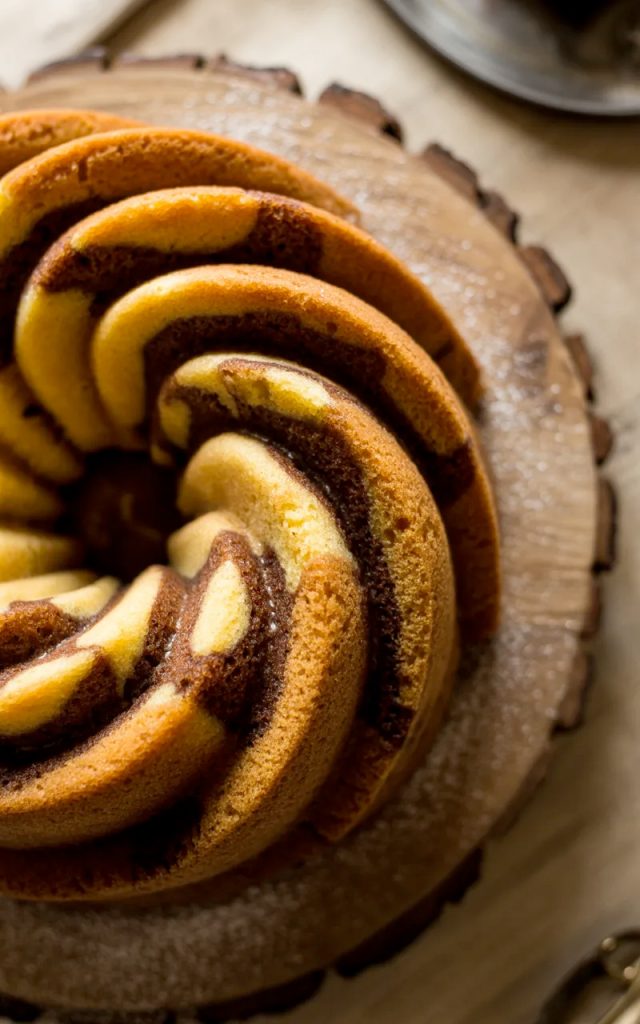 Overhead close-up of a fully baked golden cinnamon swirl bundt cake on a wooden cake stand showing the dramatic dark cinnamon sugar swirl pattern spiraling through the ridged golden cake