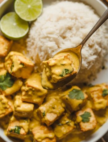 Close-up overhead shot of a white bowl filled with golden coconut lime tofu cubes in a rich creamy golden sauce with fresh cilantro fluffy white rice and lime wedges