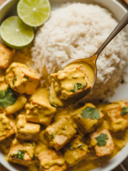 Close-up overhead shot of a white bowl filled with golden coconut lime tofu cubes in a rich creamy golden sauce with fresh cilantro fluffy white rice and lime wedges
