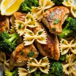Overhead close-up of a large white plate filled with cowboy butter lemon bowtie chicken with broccoli showing golden seared chicken pieces bowtie pasta and bright green broccoli florets coated in a glossy herb-flecked butter sauce with fresh parsley and lemon