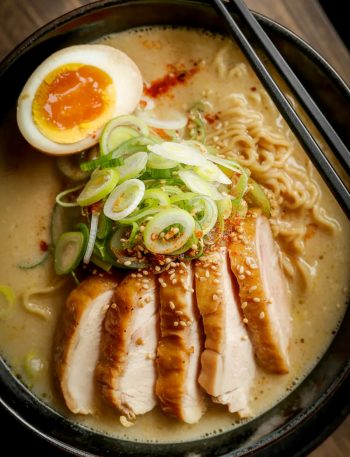 Overhead close-up of a dark bowl of creamy garlic chicken ramen with sliced seared chicken thigh soft boiled egg ramen noodles green onions chili flakes and sesame seeds in a rich golden creamy broth