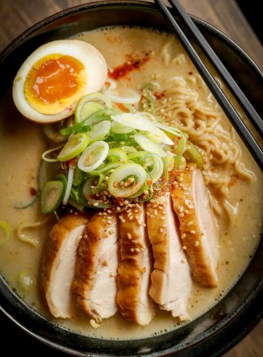 Overhead close-up of a dark bowl of creamy garlic chicken ramen with sliced seared chicken thigh soft boiled egg ramen noodles green onions chili flakes and sesame seeds in a rich golden creamy broth