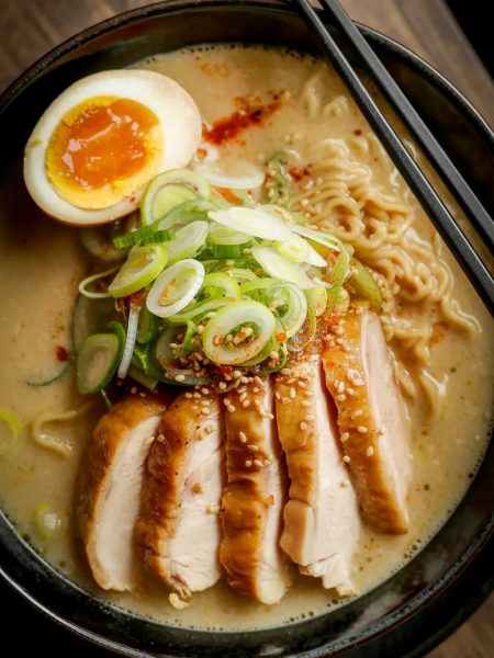 Overhead close-up of a dark bowl of creamy garlic chicken ramen with sliced seared chicken thigh soft boiled egg ramen noodles green onions chili flakes and sesame seeds in a rich golden creamy broth