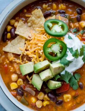 Overhead close-up of a large white bowl of creamy vegetarian tortilla soup loaded with black beans corn chickpeas and topped with tortilla chips sliced jalapenos shredded cheese and fresh avocado