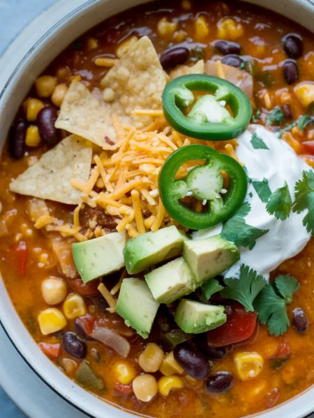 Overhead close-up of a large white bowl of creamy vegetarian tortilla soup loaded with black beans corn chickpeas and topped with tortilla chips sliced jalapenos shredded cheese and fresh avocado