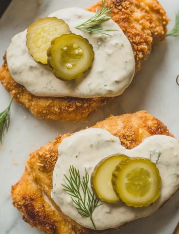 Overhead close-up of two crispy dill pickle parmesan chicken cutlets on a white marble surface topped with creamy dill pickle sauce sliced pickles and fresh dill sprigs