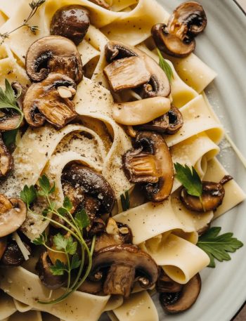 Close-up overhead shot of a large white ceramic plate piled high with garlic mushroom pappardelle pasta with golden sauteed cremini mushrooms fresh herbs and freshly grated parmesan
