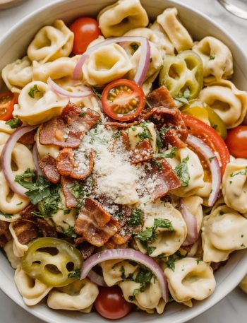 Overhead close-up of a large white bowl filled with creamy grinder tortellini salad with cheese tortellini crispy bacon cherry tomatoes red onion and freshly grated parmesan
