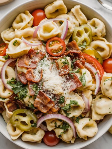 Overhead close-up of a large white bowl filled with creamy grinder tortellini salad with cheese tortellini crispy bacon cherry tomatoes red onion and freshly grated parmesan