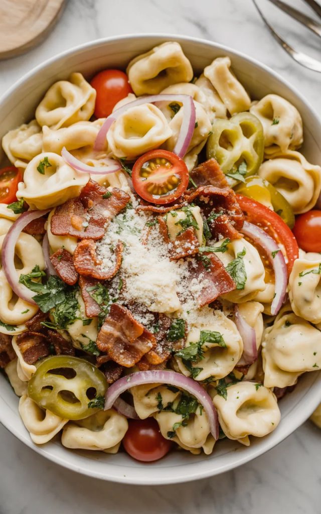 Overhead close-up of a large white bowl filled with creamy grinder tortellini salad with cheese tortellini crispy bacon cherry tomatoes red onion and freshly grated parmesan