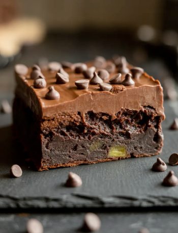 Close-up side angle of a single fudgy zucchini brownie square lifted from a slate board showing the deeply dark fudgy interior with smooth chocolate frosting and mini chocolate chips on top