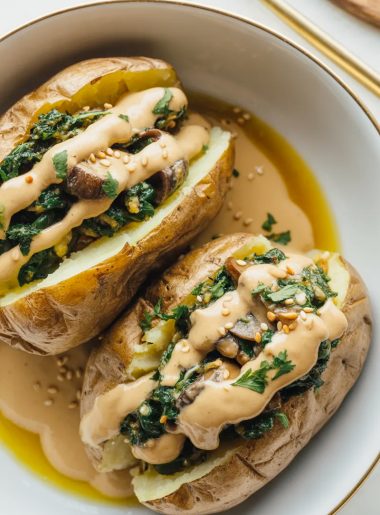 Overhead view of two mushroom and spinach stuffed baked potatoes in a white bowl drizzled with golden tahini sauce and garnished with fresh parsley and sesame seeds