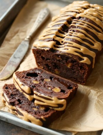 Side angle shot of a sliced peanut butter chocolate banana bread loaf on a parchment lined tray showing the dark fudgy interior with chocolate chips and a generous peanut butter drizzle across the top