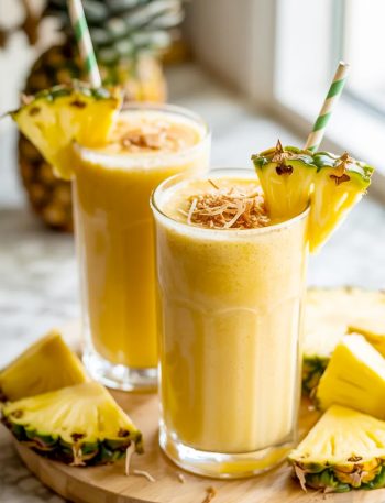 Two tall glasses of thick creamy golden pina colada smoothie garnished with fresh pineapple wedges toasted coconut and green paper straws on a wooden board surrounded by fresh pineapple pieces
