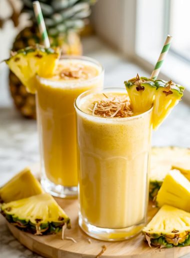 Two tall glasses of thick creamy golden pina colada smoothie garnished with fresh pineapple wedges toasted coconut and green paper straws on a wooden board surrounded by fresh pineapple pieces