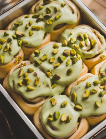 Overhead close-up of a white baking pan filled with six soft golden pistachio cream cinnamon rolls covered in smooth pale green pistachio cream cheese frosting with roughly chopped pistachios scattered generously across the top