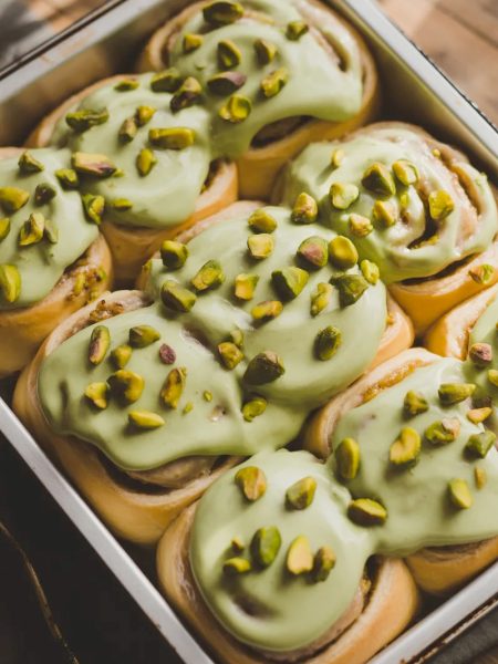 Overhead close-up of a white baking pan filled with six soft golden pistachio cream cinnamon rolls covered in smooth pale green pistachio cream cheese frosting with roughly chopped pistachios scattered generously across the top