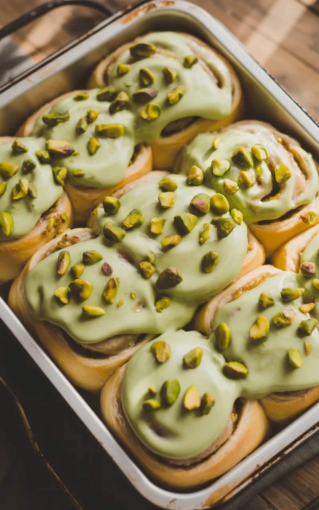 Overhead close-up of a white baking pan filled with six soft golden pistachio cream cinnamon rolls covered in smooth pale green pistachio cream cheese frosting with roughly chopped pistachios scattered generously across the top