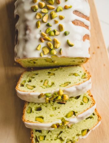 Overhead shot of a sliced pistachio pudding bread loaf on a light wooden board showing the pale green crumb with chopped pistachio pieces throughout and a white vanilla glaze drizzled across the top with chopped pistachios scattered over the glaze