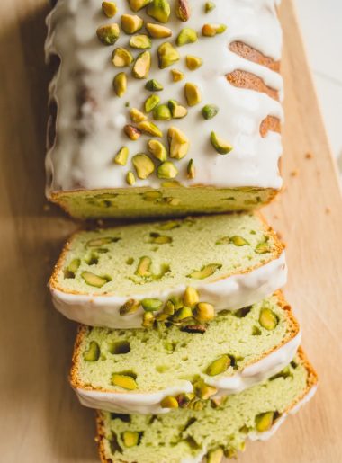 Overhead shot of a sliced pistachio pudding bread loaf on a light wooden board showing the pale green crumb with chopped pistachio pieces throughout and a white vanilla glaze drizzled across the top with chopped pistachios scattered over the glaze
