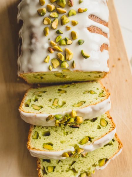 Overhead shot of a sliced pistachio pudding bread loaf on a light wooden board showing the pale green crumb with chopped pistachio pieces throughout and a white vanilla glaze drizzled across the top with chopped pistachios scattered over the glaze