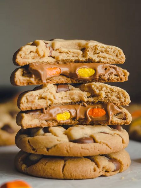 Close-up dramatic shot of a stack of three Reese's Pieces peanut butter cookies with the top cookie broken in half showing the soft chewy interior with melted Reese's Pieces candy throughout