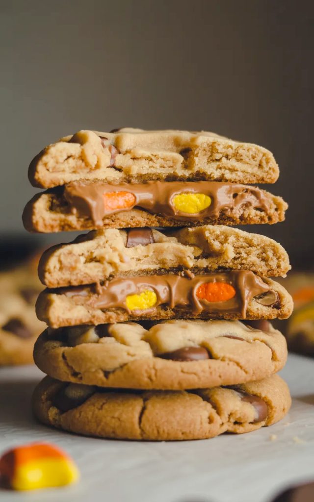 Close-up dramatic shot of a stack of three Reese's Pieces peanut butter cookies with the top cookie broken in half showing the soft chewy interior with melted Reese's Pieces candy throughout