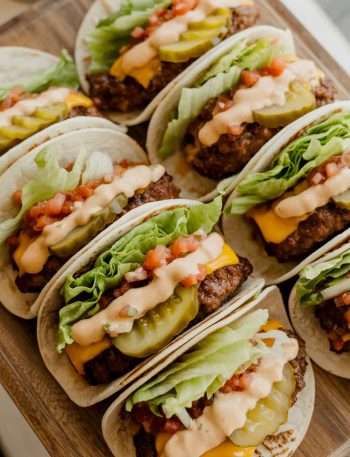 Six smash burger tacos on a wooden serving board loaded with melted cheddar, shredded lettuce, diced tomatoes, pickles, and special sauce drizzle